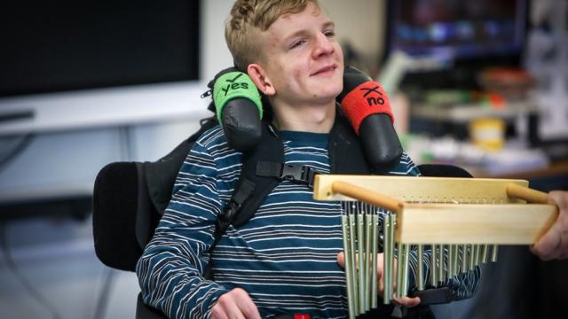 Treloar College student playing an instrument by touching it; his assistant is holding the instrument.