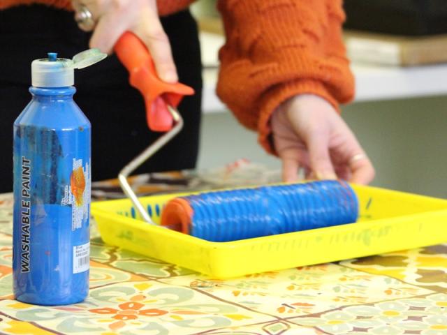 A yellow tray with blue paint in and a bottle of blue paint next to it: Rachel the art therapist is using a roller to dip in the paint.