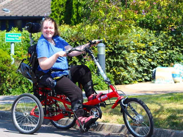 Treloar's student is using a trike; she is smiling at the camera and squinting as it's a very sunny day; green shrubs and trees in the background.