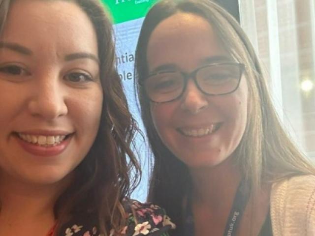 Claire taking a selfie with a woman  at the conference; A screen with a presentation about Treloar's in the background.