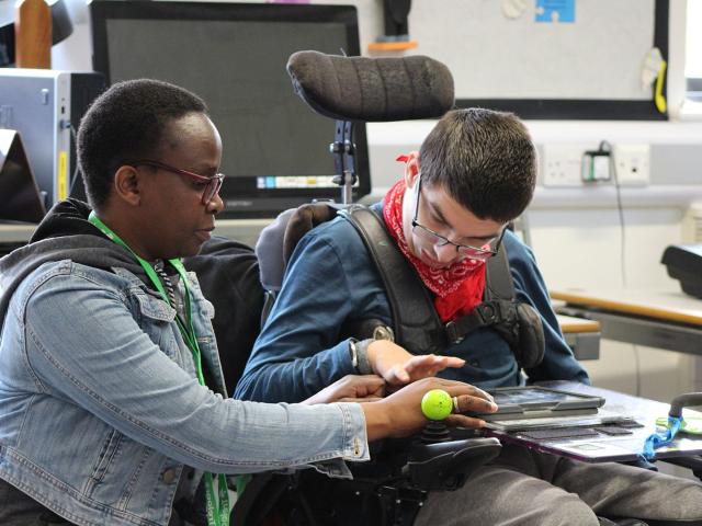 Treloar College using his iPad to play clarion; his assistant is helping him by supporting his hand.