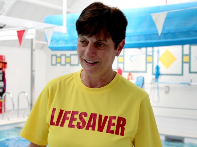 Kim Smith, Sports Coach and Event Leader at Treloar's pictured wearing bright yellow T-shirt with a red writing on her chest saying Lifesaver; in the background, there's a swimming pool area.