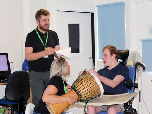 Tom Clarke in the classroom teaching his class: he is standing in front of one student who is playing a big drum - the drum is being held by his assistant.