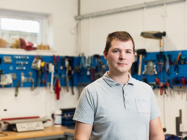 Portrait of Dean Hall, an Assistive Technologist at Treloar’s. In the background, multiple tools hanging on the walls of the workshop. 