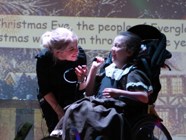 Donna, Deputy Lead Student Support Assistant at Treloar’s, on the stage with Treloar School student during a Christmas performance: she is holding a microphone helping student sing; both are smiling and having a good time.