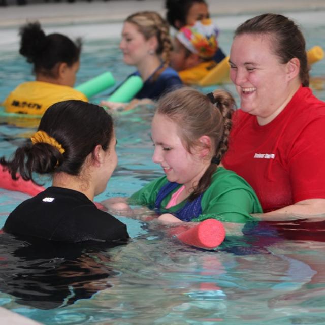 Treloar School student in the pool, supported by 2 members of staff holding onto a pink swimming noodle; in the background other students and staff.