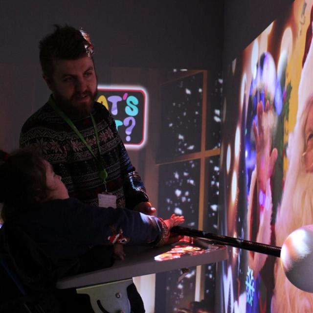 An interactive projections on the wall of Santa, accompanied by scents and sounds: Treloar College student is being assisted by her teacher to interactive with the projection by holding a big pole with a round end.