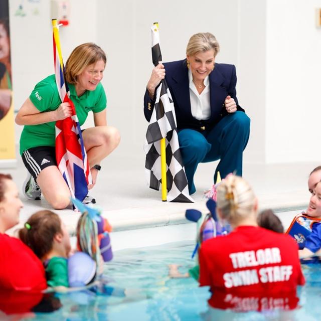 Her Royal Highness The Duchess of Edinburgh talking to Treloar's students who are in the pool with their assistants. The students have just finished a race and Her Royal Highness is holding a chequered flag.