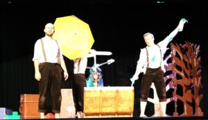 3 performers from Box Tale Soup on stage at Treloar's: one is holding a big yellow umbrella, one is holding a bunting and one is standing empty-handed