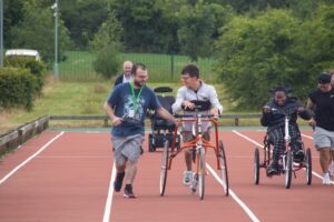 Treloar College student taking part in the annual Sports Day: he is using 3 wheeled walking frame supported by 2 members of staff. 