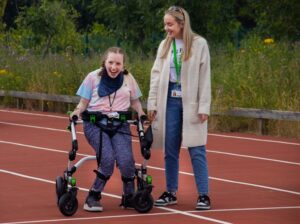 Treloar College student taking part in the annual Sports Day: she is using her walking frame, supported by her assistant; she's on the athletic track, smiling.
